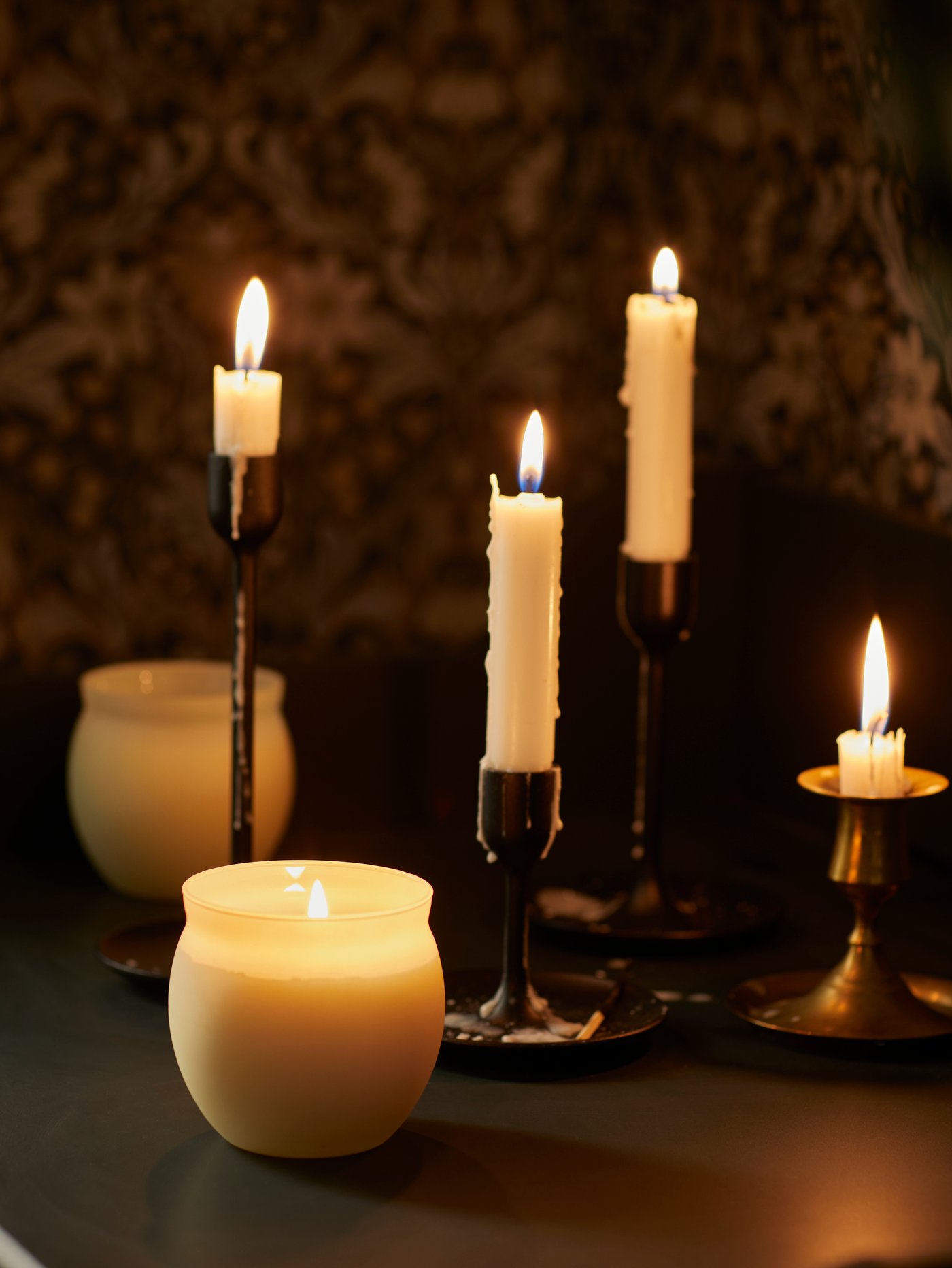 Un salón moderno con una vela perfumada en un vaso y un juego de tres portavelas negros con velas encendidas.