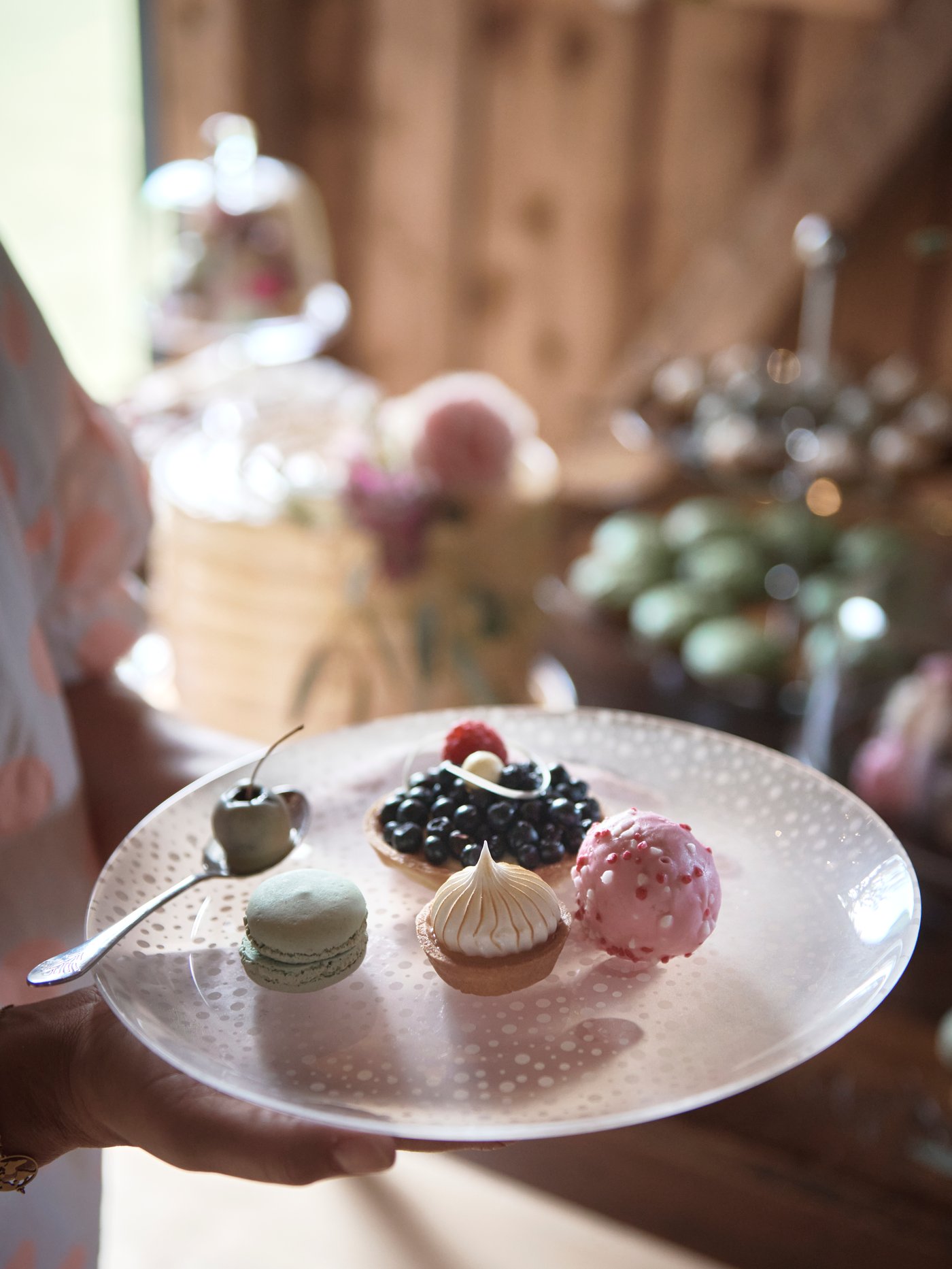 A woman at a party holds in her hands a frosted glass INBJUDEN plate laden with several mini pastries and sweets.