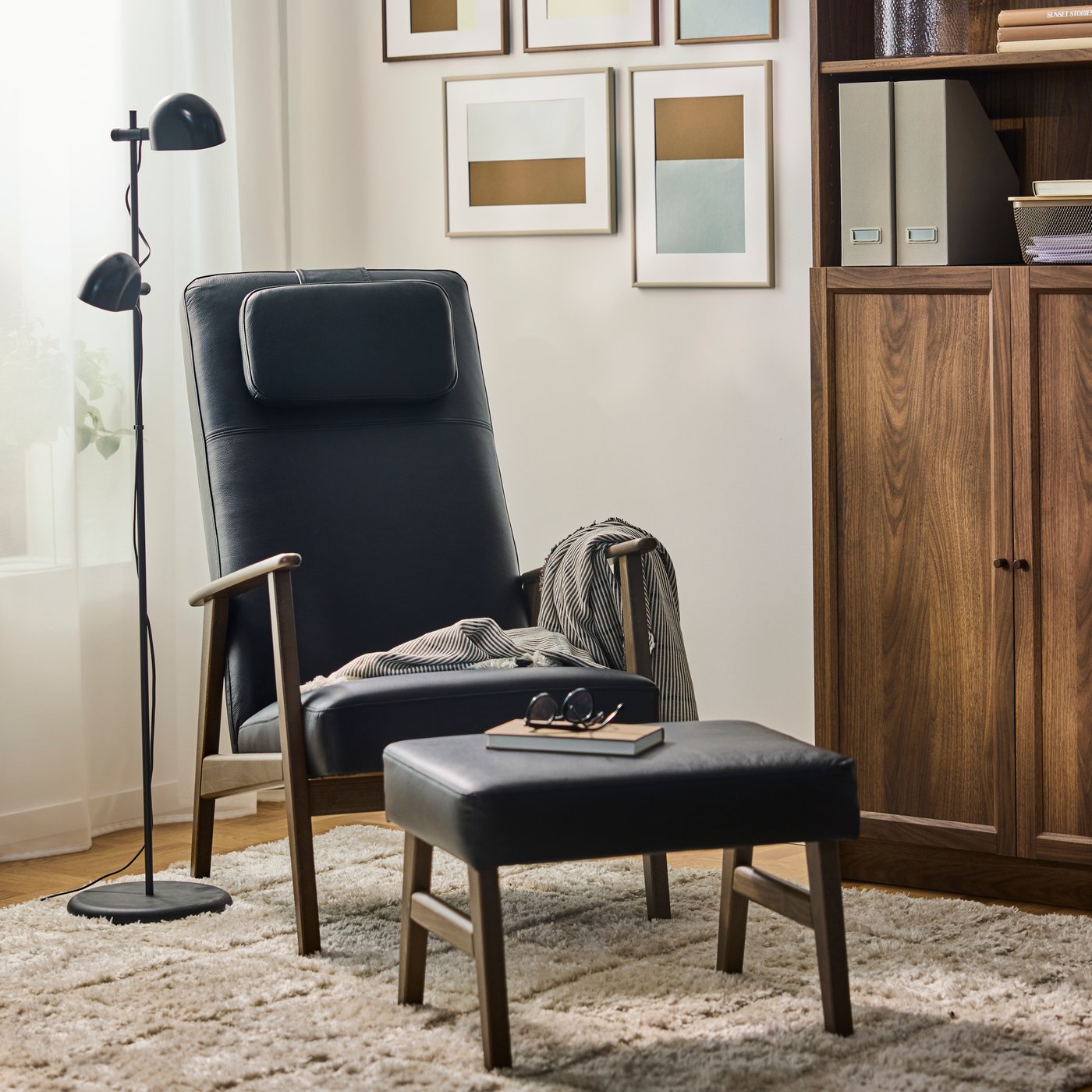 An EKENÄSET high-back armchair and footstool with a wooden frames and a dark grey throw and beside a floor lamp and cabinet.