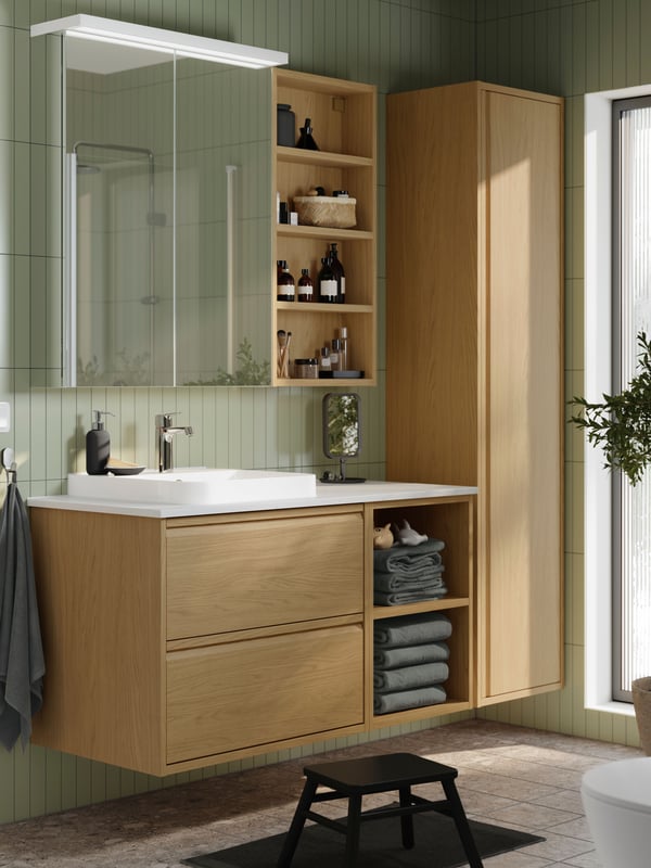 A green-tiled bathroom has an ÄNGSJÖN vanity sink in oak effect with an open frame for grey towels and a tall cabinet.
