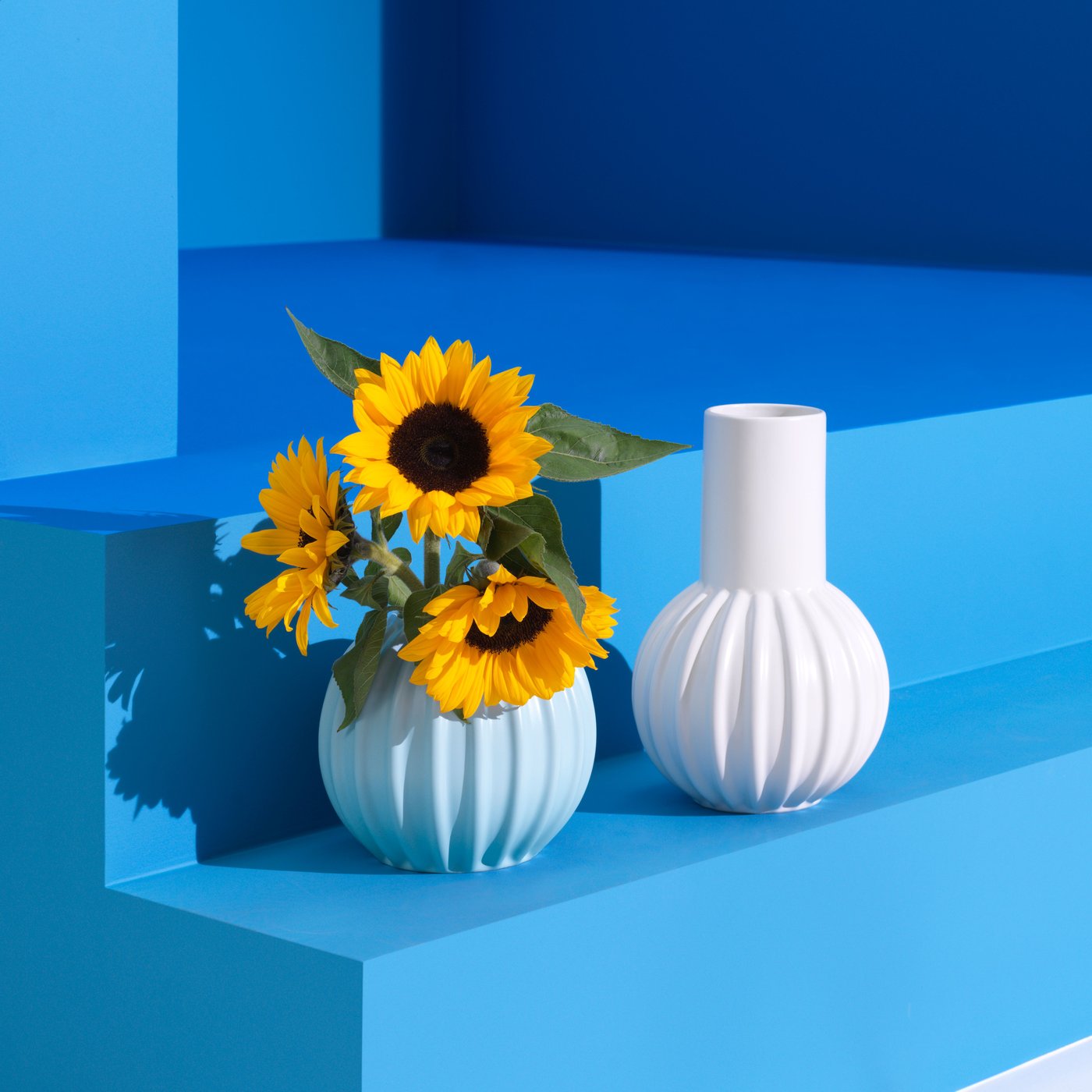 Devant un mur bleu, tournesols jaunes dans un vase SKOGSTUNDRA bleu clair.