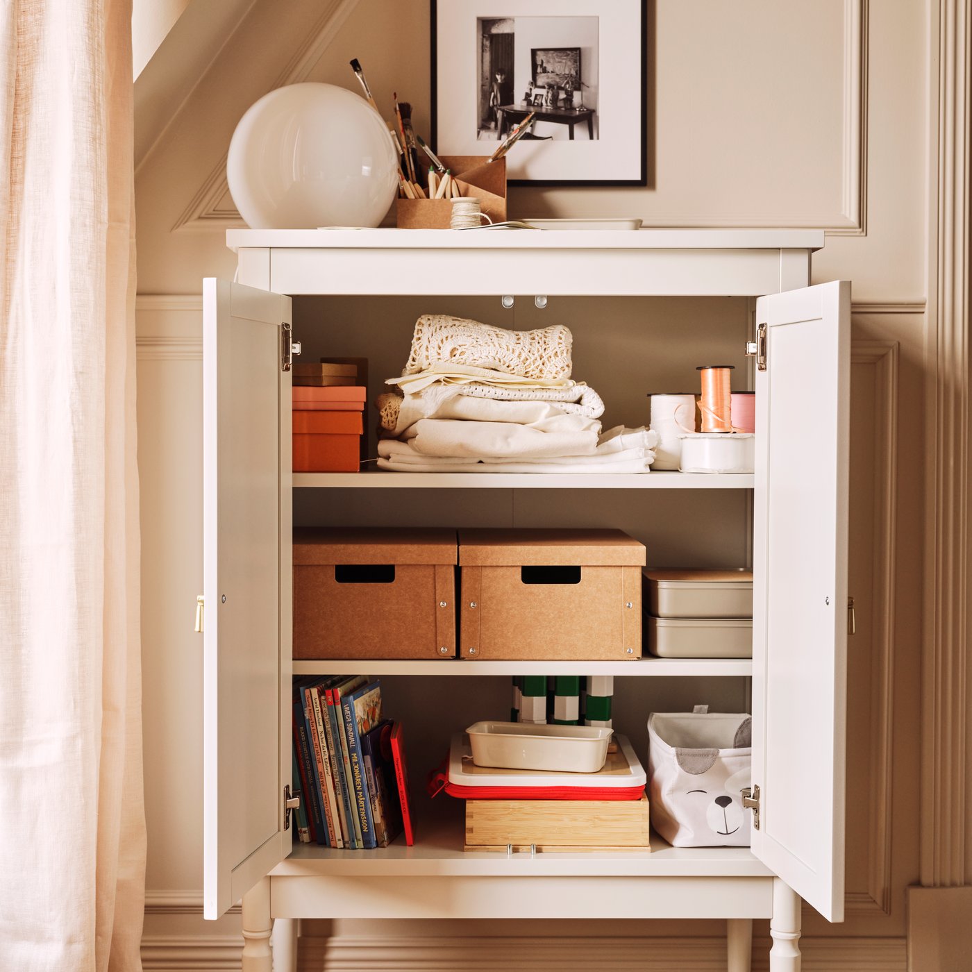 Kremowa szafka FÄRJKARL z półkami, na których poukładane są różne przedmioty, w tym dwa pojemniki GÄRDESGÅRD oraz pudełko DRÖMSLOTT.