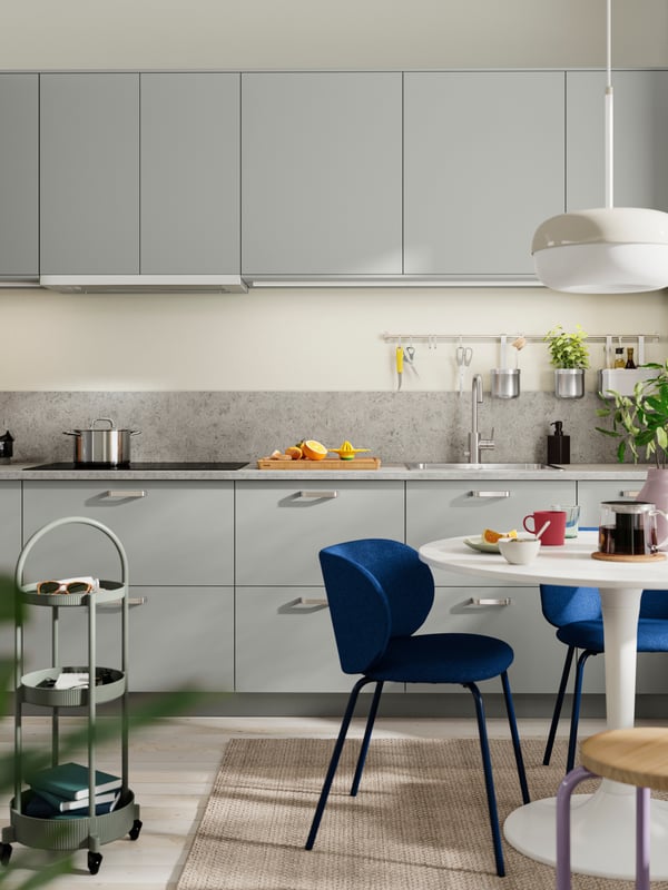 A kitchen with HAVSTORP light grey fronts with silver handles, a dining table with blue chairs, and a trolley beside a rug.