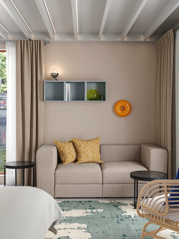 A hotel room with a JÄTTEBO 2-seat modular sofa, cushions, coffee tables and a round, orange VARMBLIXT LED table/wall lamp.