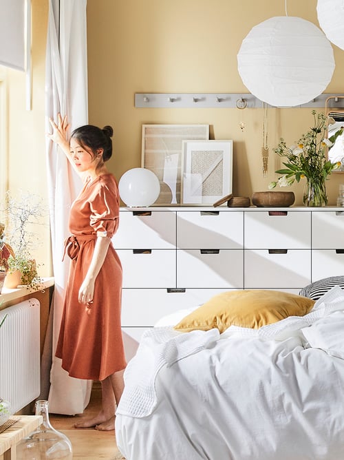 A woman in a white and yellow bedroom in front of a white NORDLI combination of drawers with pictures and baskets on top.