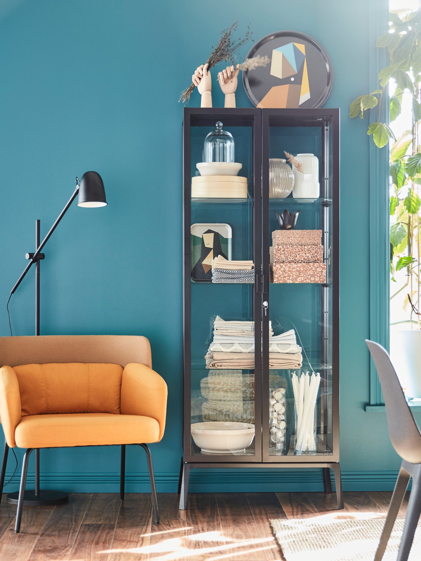 Comedor con una vitrina gris con cajas y dos manos decorativas, una lámpara de lectura de pie y un sillón amarillo.