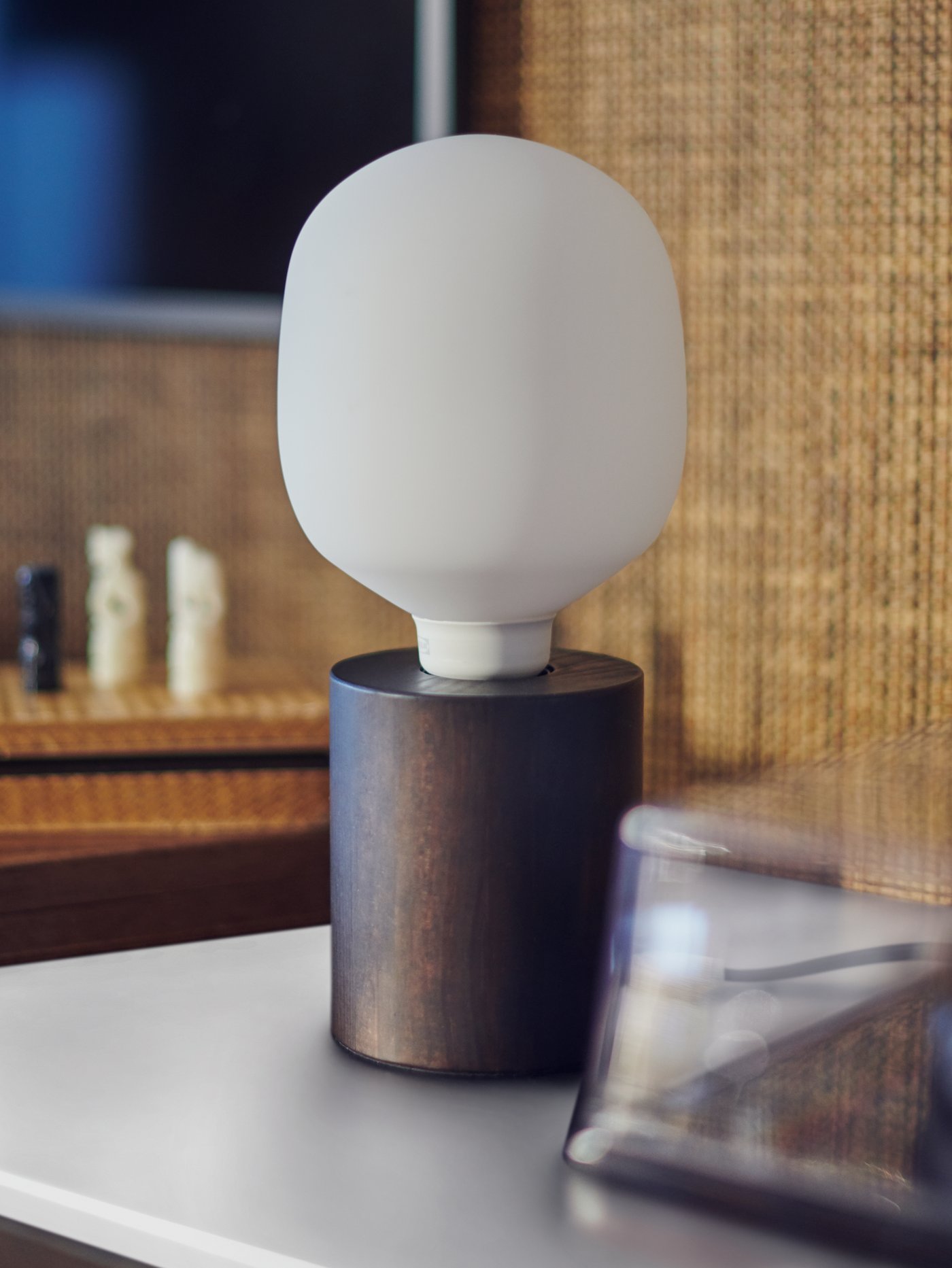 A BLEKKLINT table lamp with a dark brown wood base on a white surface in front of a wall covered in natural seagrass.