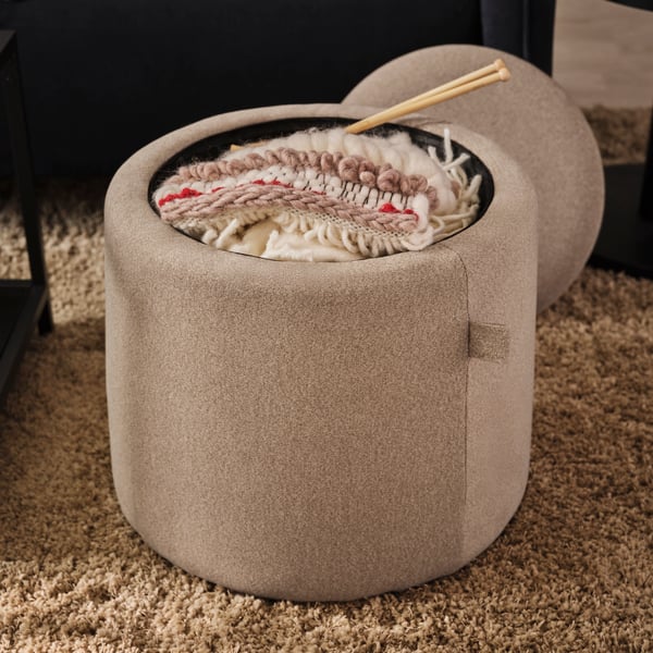 A dark-beige JÄRRESTAD pouffe, its top off to reveal a knitting kit in the storage compartment, standing on a high-pile rug.