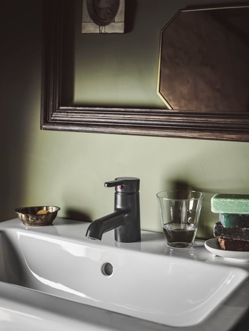 Close-up of a black wash basin mixer tap fixed to a white, single wash basin. A glass with water is next to the mixer tap. 