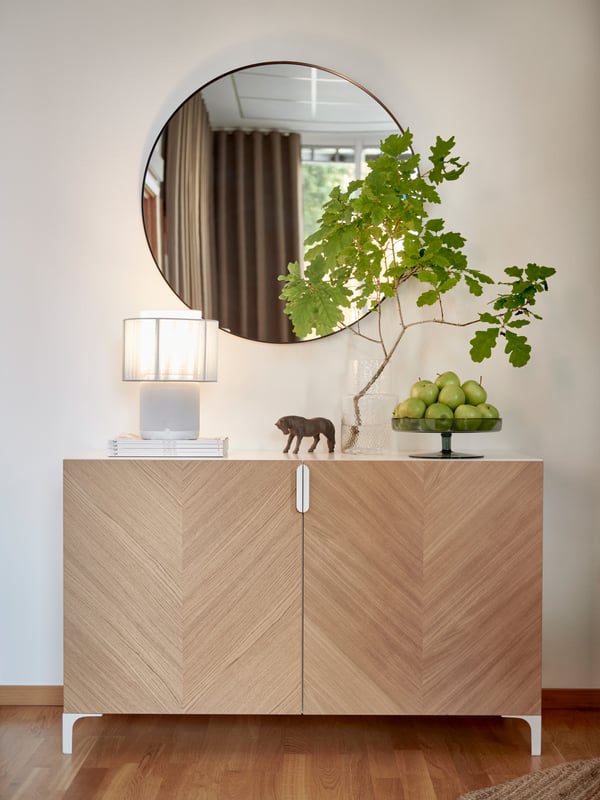 Ett HEDEVIKEN träskåp i chevronmönster med vit toppyta står mot en vit vägg. En bordslampa med vit skärm, en dekorativ figur, fat med gröna äpplen och en glasvas med gröna kvistar. Rund spegel ovanför skåpet reflekterar gardiner och fönster.
