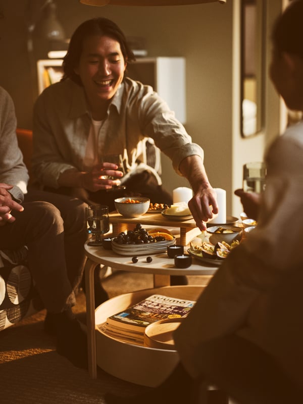 A person extends their hand over a STOLTHET chopping board with GLADELIG bowl, and plates, all set on a BORGEBY coffee table.