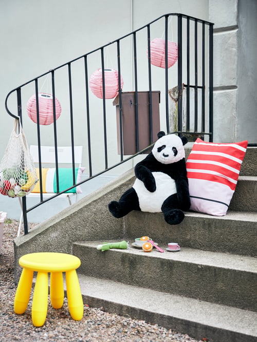 Cadru de exterior cu scări, taburet galben pentru copii, lămpi roz cu pandantiv, jucărie moale panda, scaun alb și perne cu dungi.