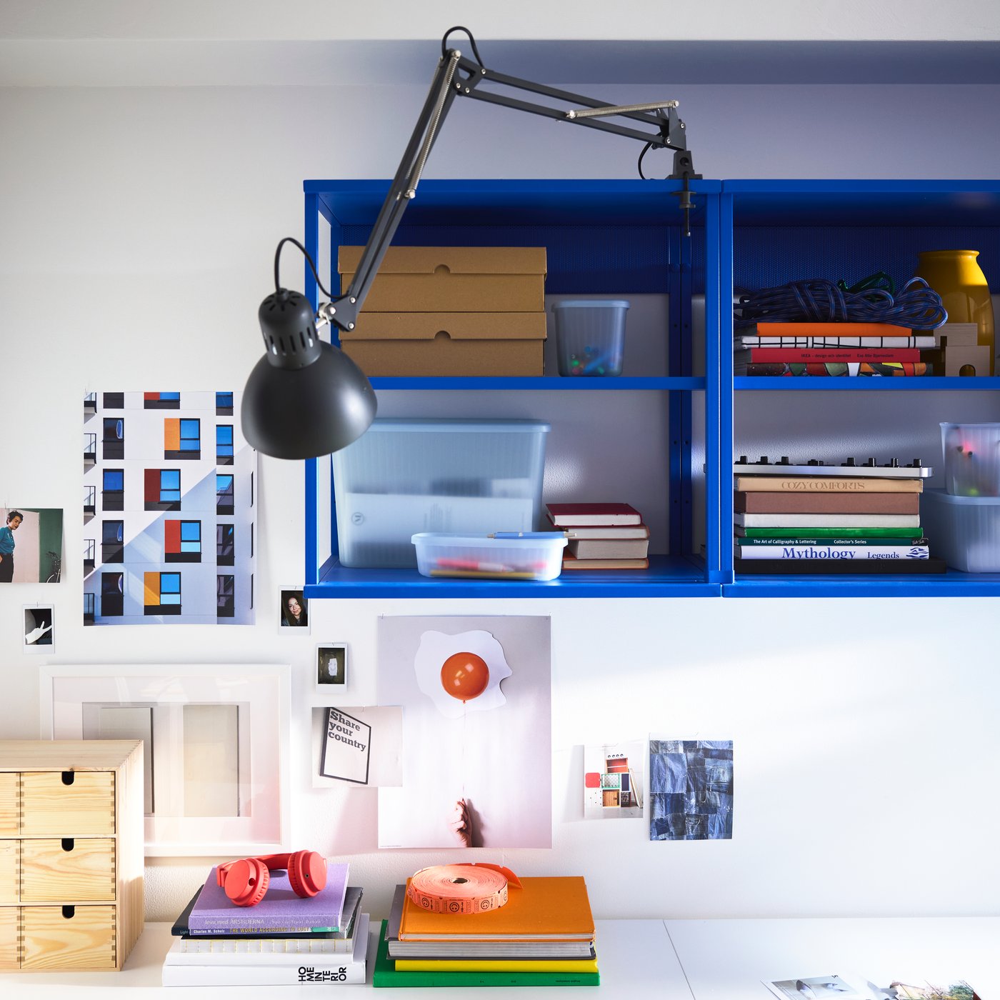 A bright blue shelving unit on the wall on top of desk, a black TERTIAL work lamp, piles of books and things on the desk.