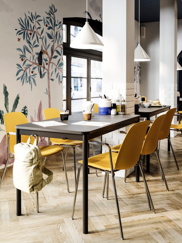 Une table à manger avec des crochets sur le côté où un sac beige est suspendu, des chaises jaune foncé, des suspensions blanches et un mur peint.