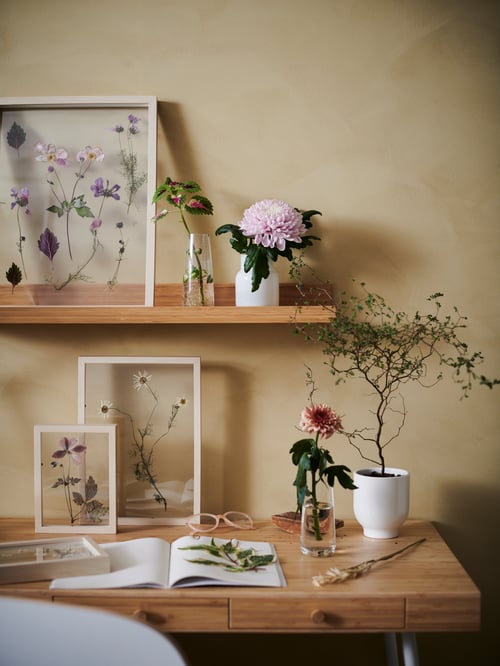 A potted plant and flowers in vases and frames fill a desk and a MÅLERÅS bamboo picture ledge on the wall above.