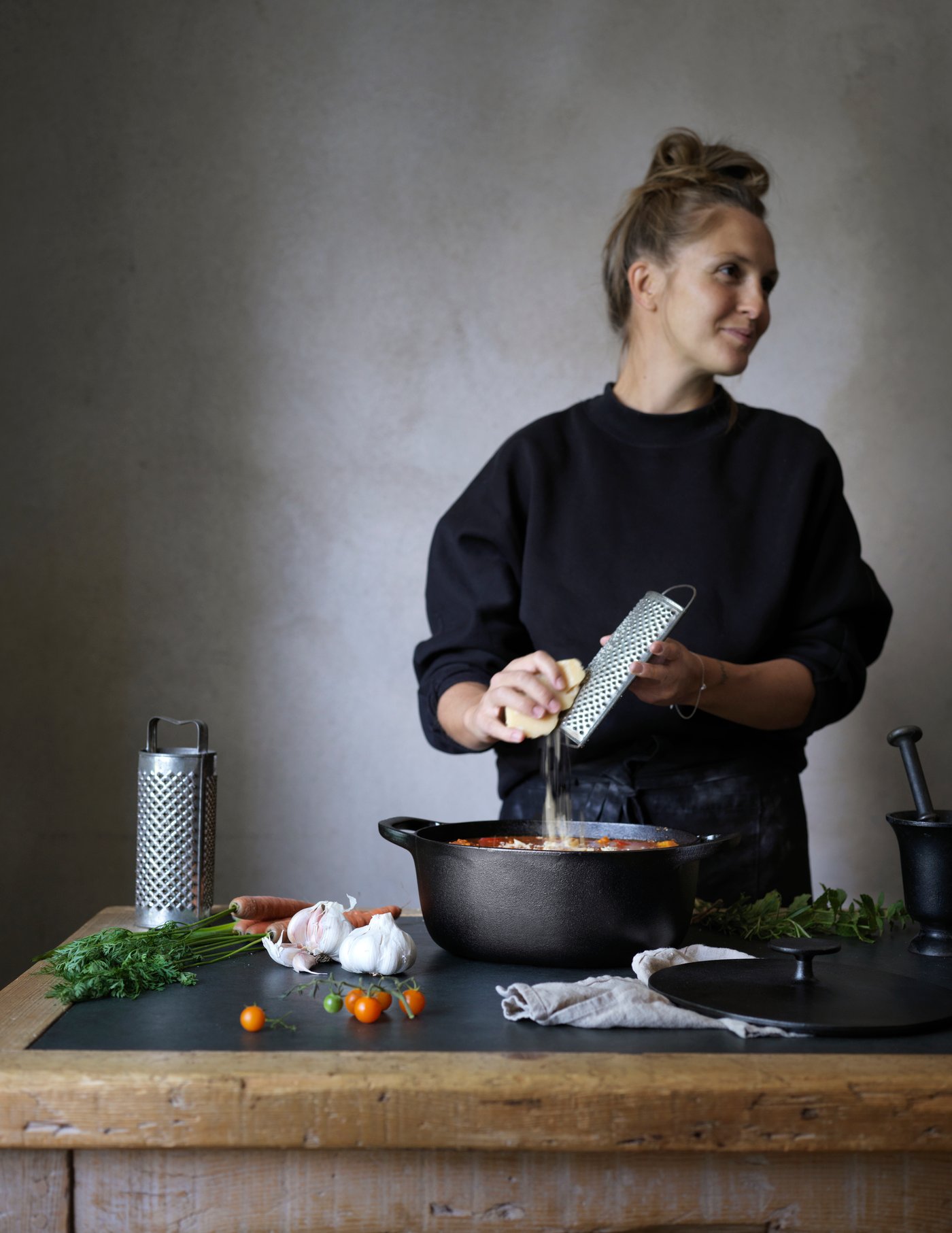 En kvinde river ost ned i en gryde fyldt med mad omkring gryden ligger forskellige ingredienser og yderst til venstre står et rivejern op.