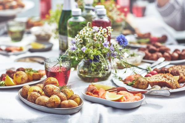 Et bord dækket med forskellige midsommer retter serveret på grå STOCKHOLM 2025 serveringsfade, som laks, kartofler, brød og planteboller samt en vase med friske blomster.