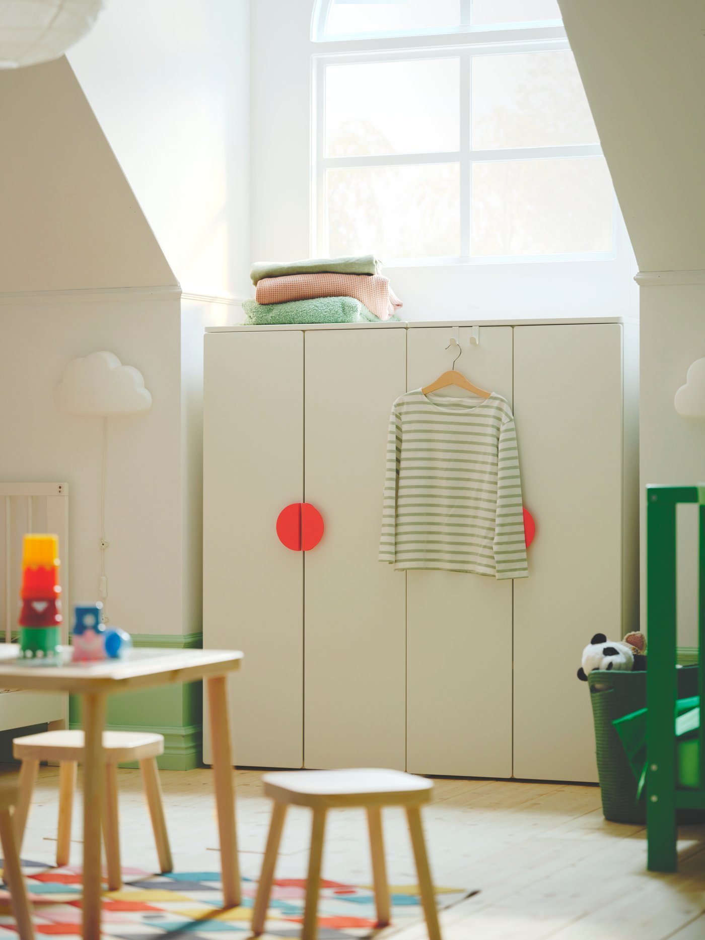 Ett vitt SMÅSTAD garderobsskåp med ljusröda BEGRIPA handtag står framför ett fönster. En vit UPPLYST LED vägglampa sitter bredvid.

