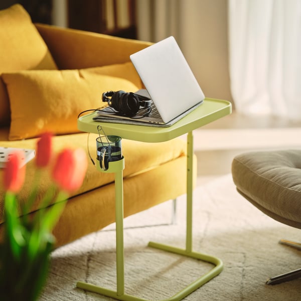 A living room with a BJÖRKÅSEN laptop stand displaying a laptop and headphones near a yellow sofa.