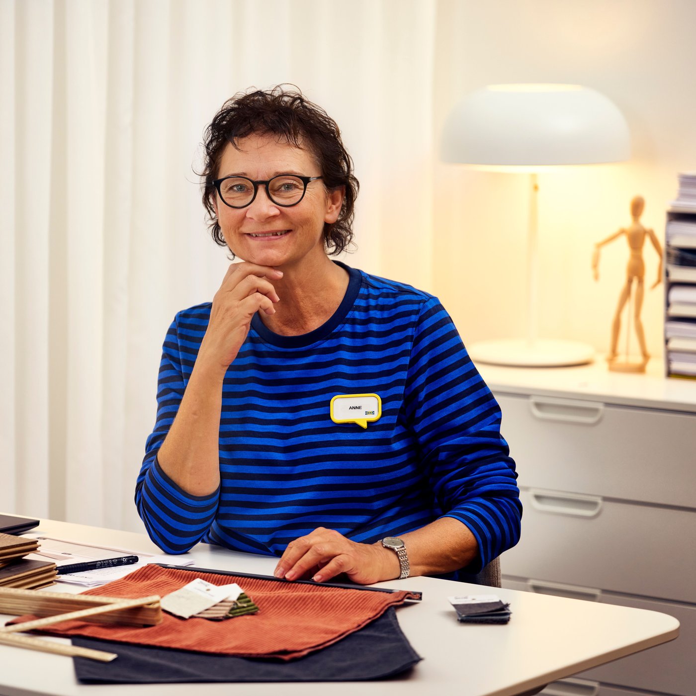 En IKEA-indretningsarkitekt iført en blå sweater sidder ved et skrivebord med indretningsprøver og smiler til kameraet.