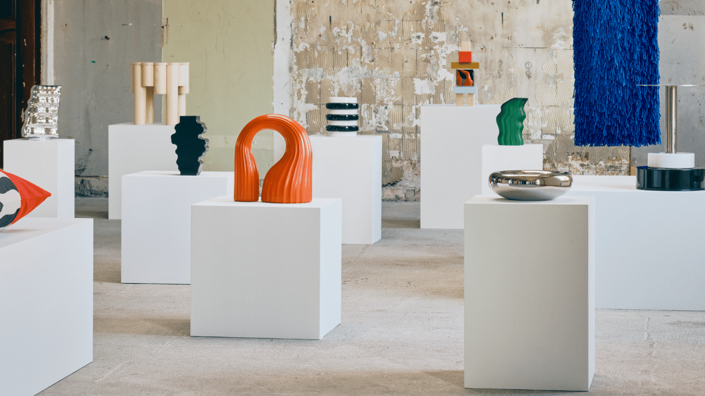 Galerie contemporană într-un spațiu industrial uzat, cu sculpturi abstracte pe piedestale albe: o buclă portocalie, un bol metalic reflectorizant, forme ondulate verzi și negre și o piesă textilă albastră agățată.