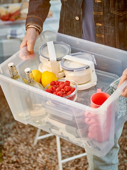 Eine Person hält eine transparente SAMLA Box in den Händen, die mit Geschirr, Glasflaschen und Behältern gefüllt ist, um die Kühlakkus gebunden sind.