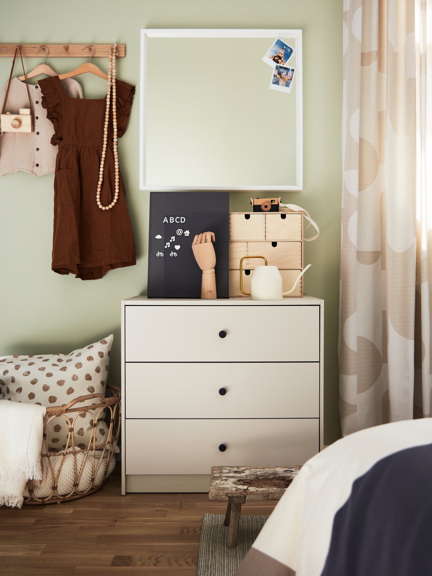 Habitación para bebés y niños con una cómoda beige de 3 cajones de madera, entre un par de cortinas oscurecedoras y una cesta.