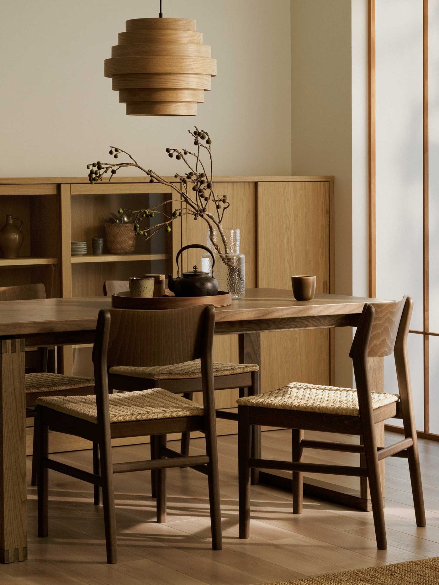 A dining room featuring a long, rectangular wooden dining table with four matching wooden chairs that have woven seats. Above the table hangs a unique, layered wooden pendant light. In the background, there's a light-coloured wooden cabinet with open shelves displaying various decorative items and closed sections. To the right, a tall screen with translucent panels allows soft light to filter in. A branch with dark berries in a vase sits on the table, along with a dark teapot and small cups on a wooden tray, and a single brown mug. The room has a warm, natural aesthetic with light-coloured walls and wooden flooring.