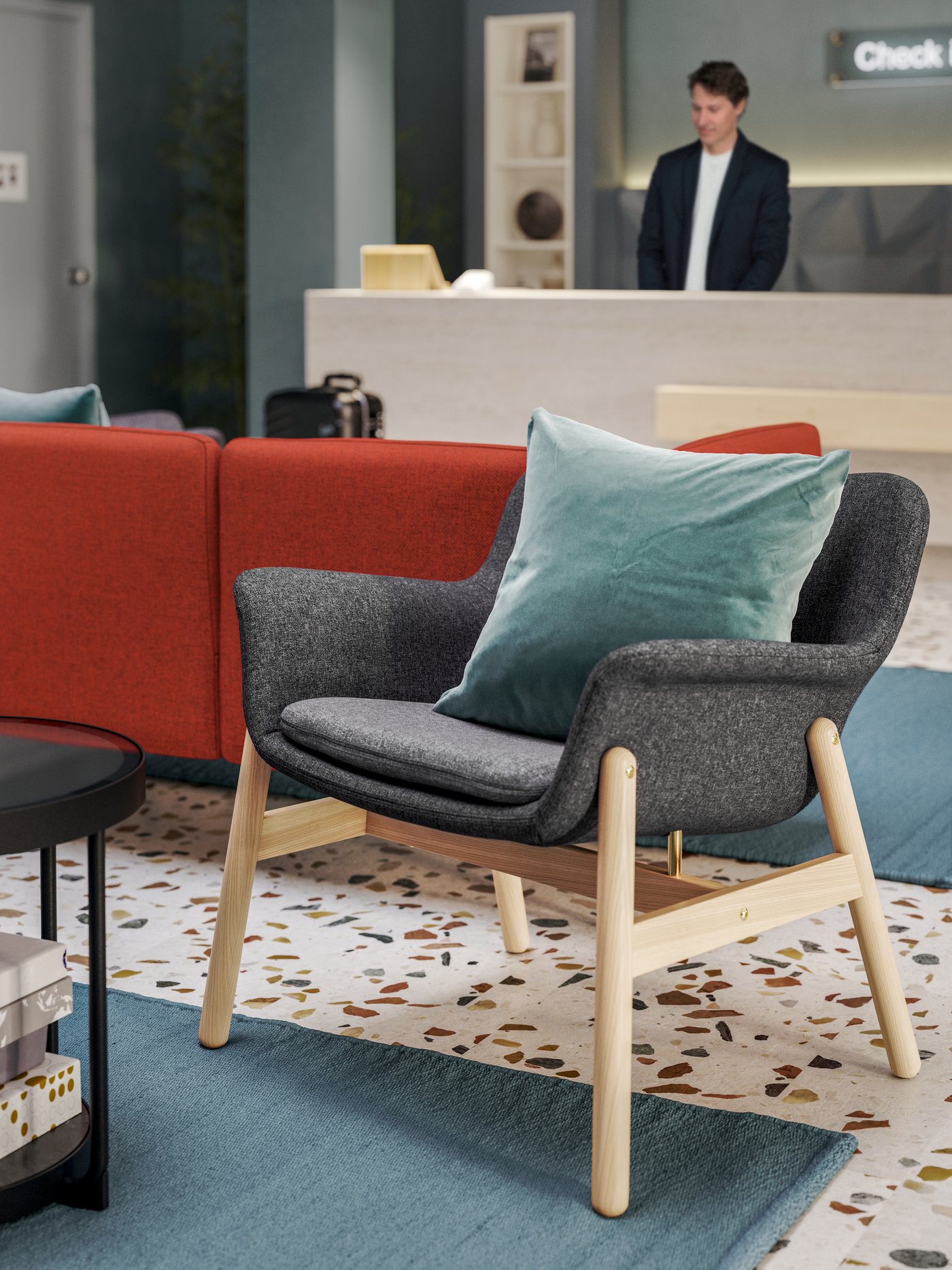  كرسي بذراعين VEDBO رمادي غامق بأرجل من خشب بتولا وراء كنبة حمراء تواجه مكتب استقبال فندق يجلس عليه موظف.