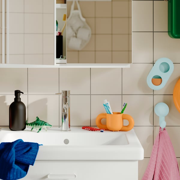 An orange HUMLESJÖN mug containing toothbrushes on a bathroom counter beside a sink and soap dispenser.