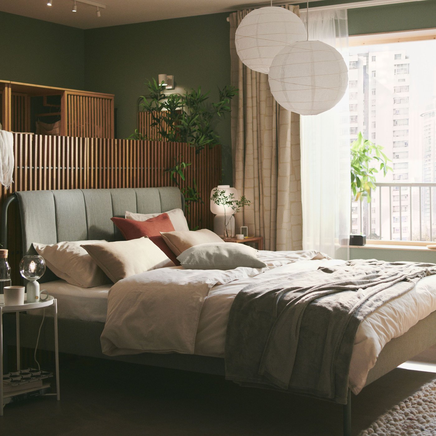 Ein gepolstertes TÄLLÅSEN Bettgestell in Graugrün mit einem Sichtschutz hinter dem Bettgestell steht in einem ruhigen grünen Schlafzimmer mit grossen Fenstern.