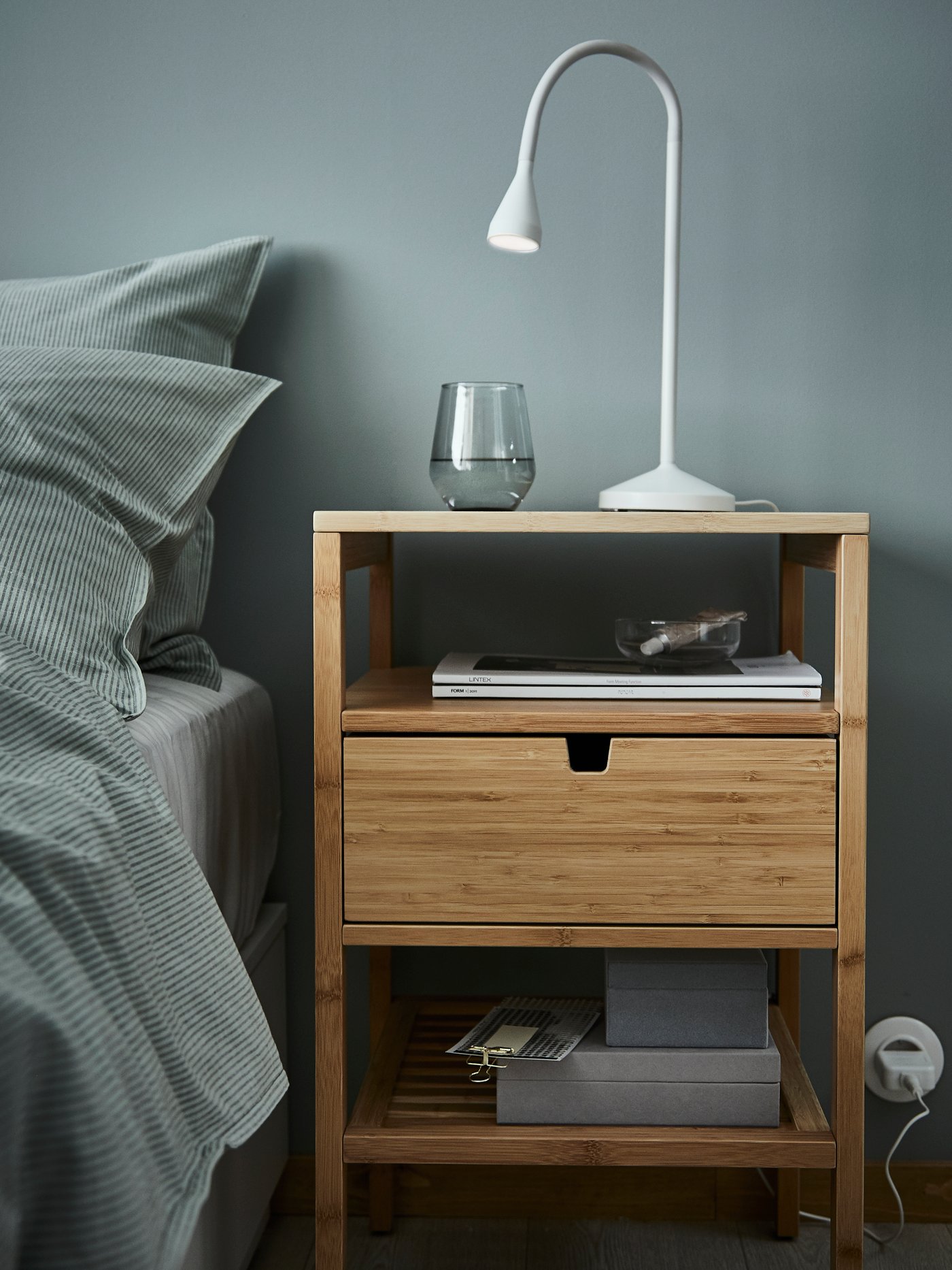Dormitorio con mesilla de noche de madera, funda nórdica y dos fundas de almohada, estructura de cama, lámpara de trabajo LED, vidrio y cajas de almacenaje con tapa.