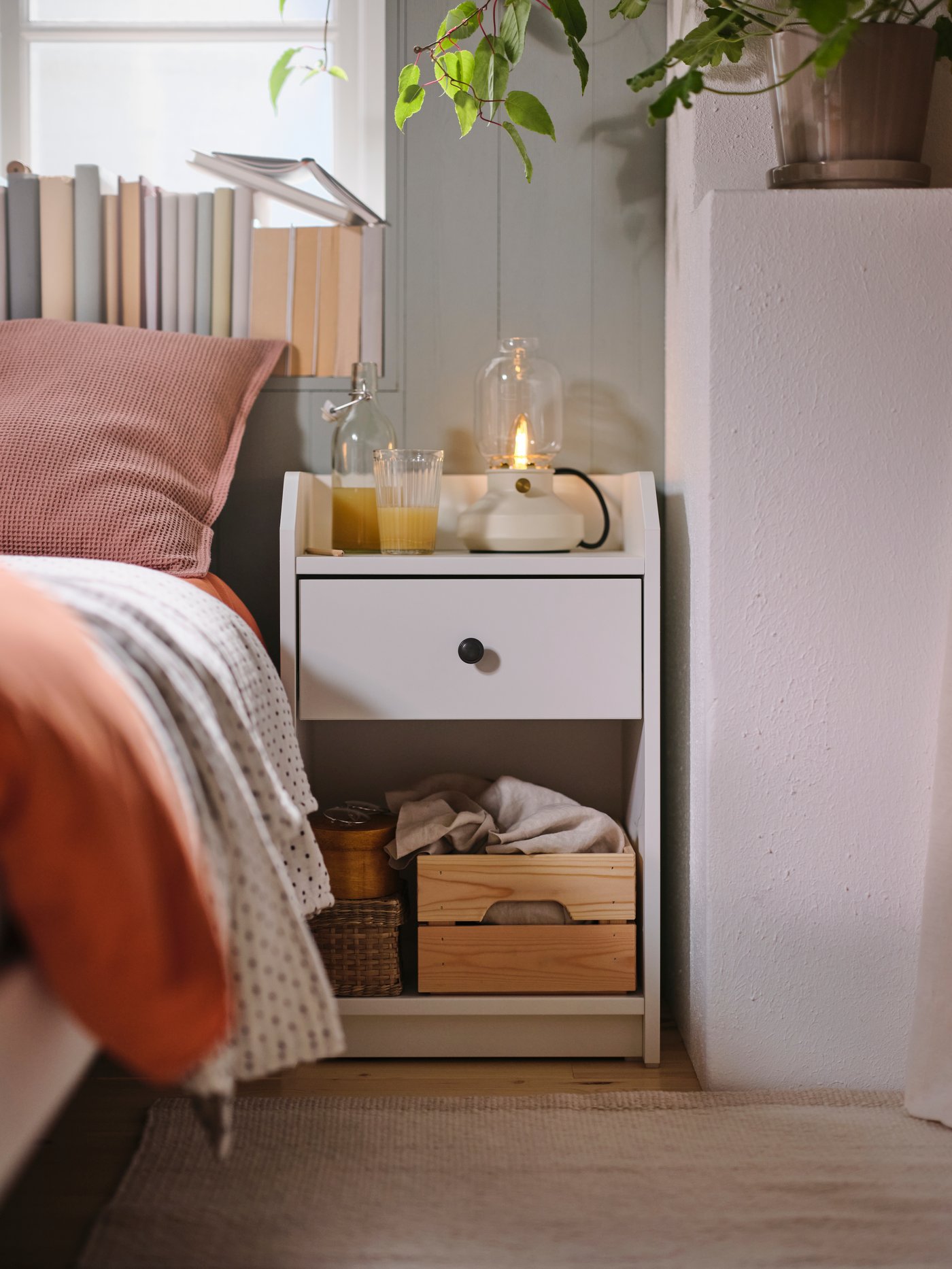 A white HAUGA bedside table with a TÄRNBY table lamp on top. A cushion in a pale pink KLOTSTARR cover is on the bed by it.