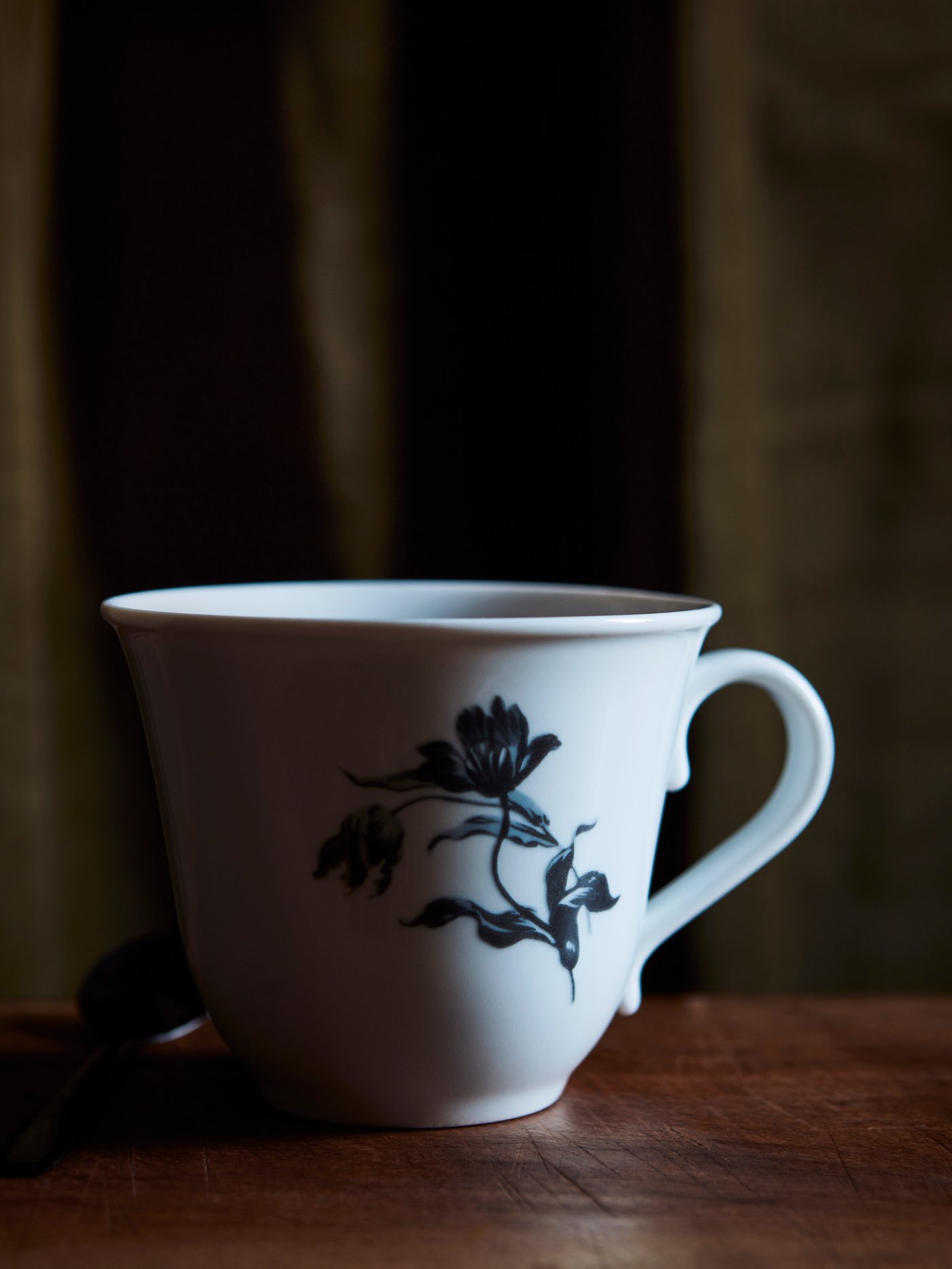 A close-up of UPPLAGA mug with a rustic floral pattern on white placed in a black room with shadows.
