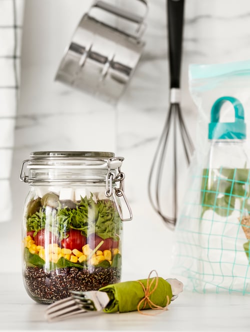 Close up on white kitchen counter with glass jar with lid containing a salad, packed cutlery, kitchen utensils on the wall.