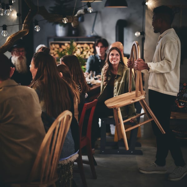A young man brings a wooden dining chair to a long table surrounded by seated dinner guests.