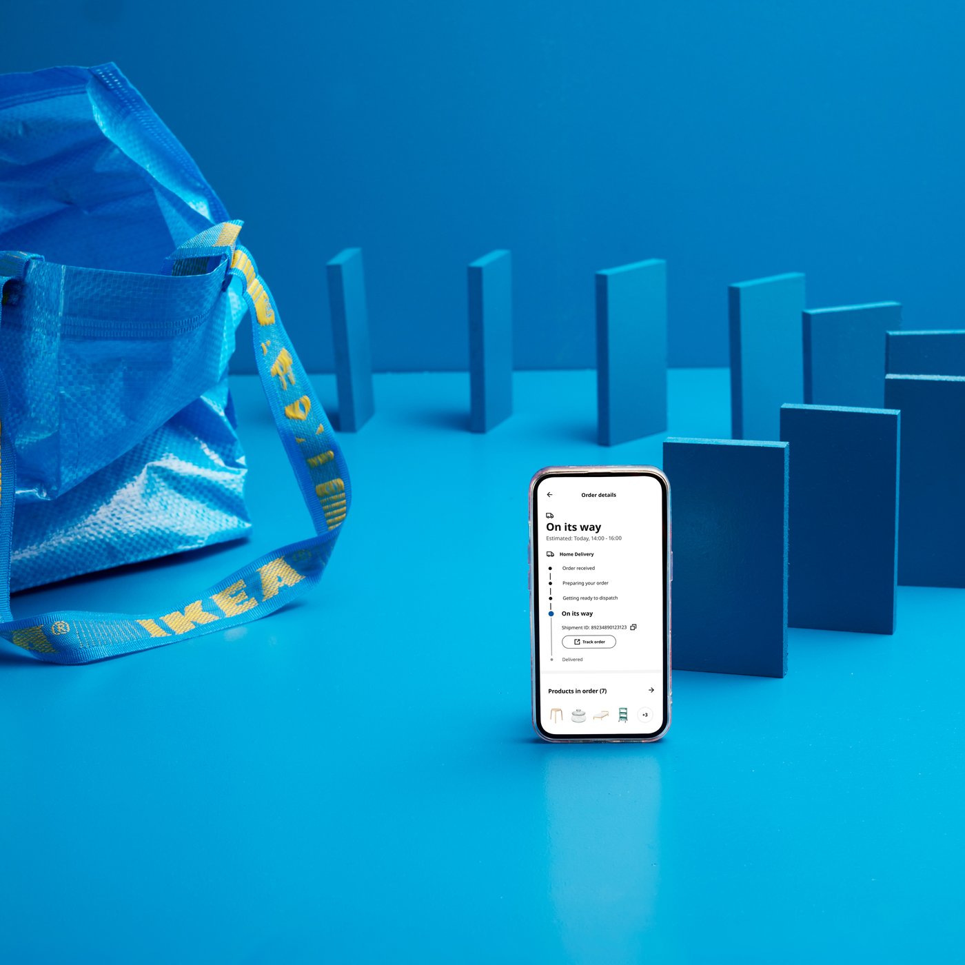 A smartphone stands upright on a bright blue surface displaying an IKEA delivery tracking screen, with blue domino-like shapes in the background.