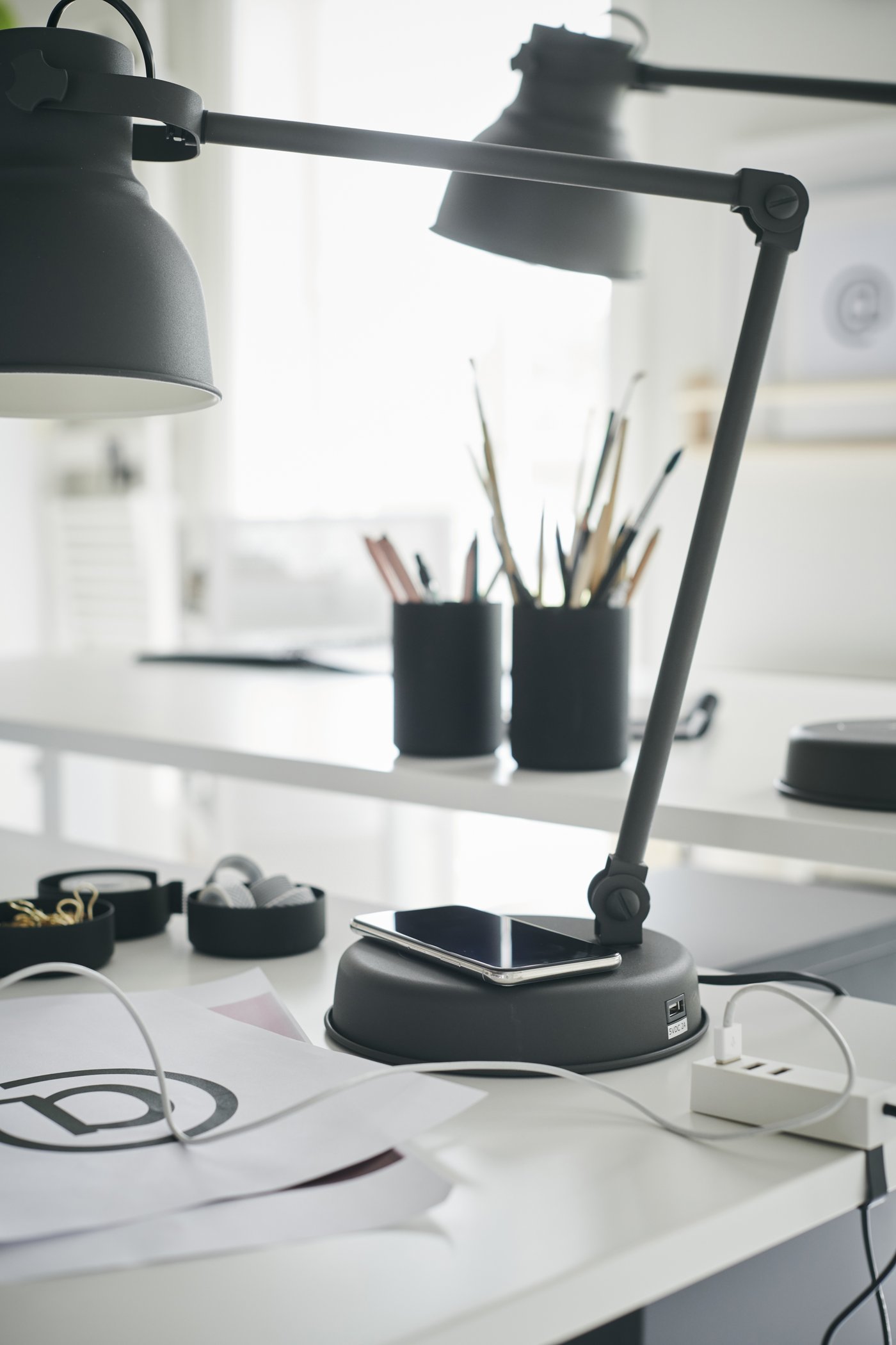 Ein Smartphone auf HEKTAR Arbeitsleuchte mit Ladefunktion in Dunkelgrau, die auf einem weißen Schreibtisch steht.