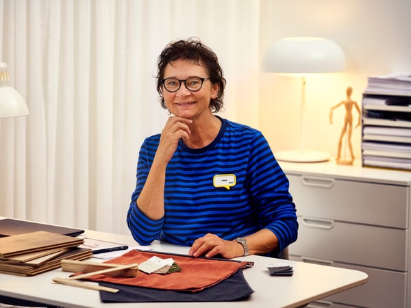 An IKEA interior designer wearing a blue sweater is sitting by a desk with interior samples, smiling to the camera.