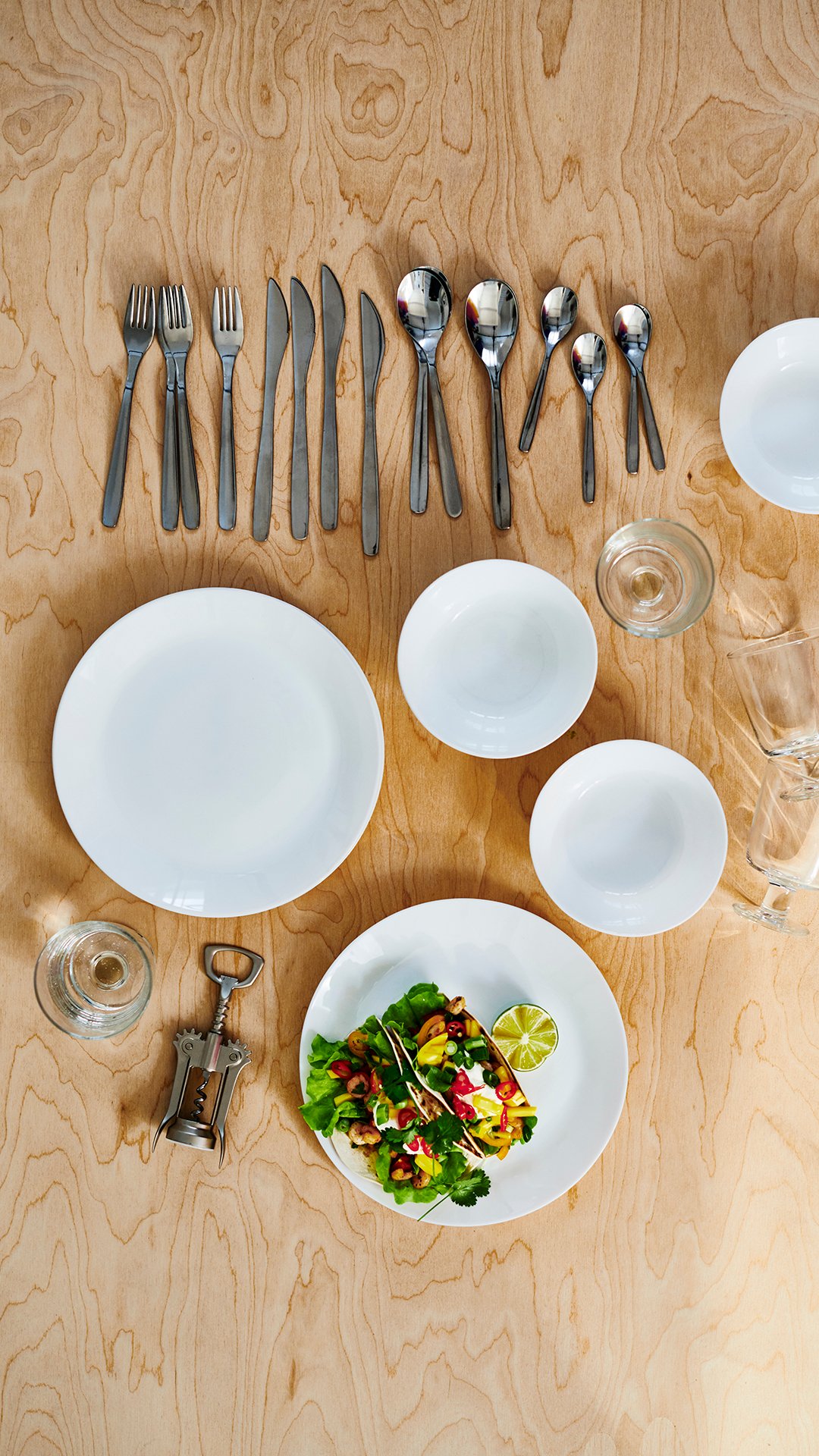 Porcelæn og bestik på en lys træoverflade: Hvide OFTAST tallerkener og skåle, MOPSIG bestik, vinglas og proptrækker.