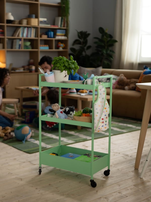 Un carrello portatutto NISSAFORS verde chiaro a tre ripiani, che contiene giochi per bambini, una piantina e altri oggetti di uso quotidiano.
