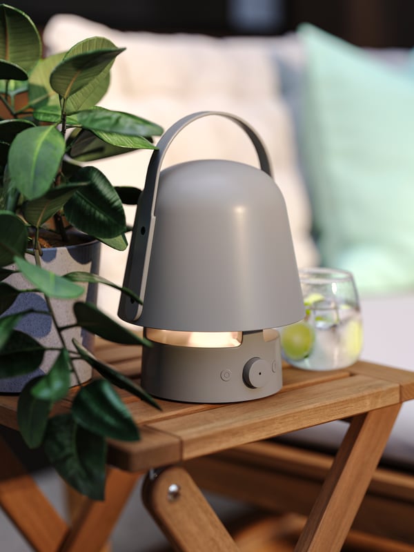 O zonă de relaxare amenajată pe o terasă, cu o lampă cu boxă Bluetooth VAPPEBY de exterior gri pe un taburet NÄMMARÖ din lemn de acacia lângă o plantă.