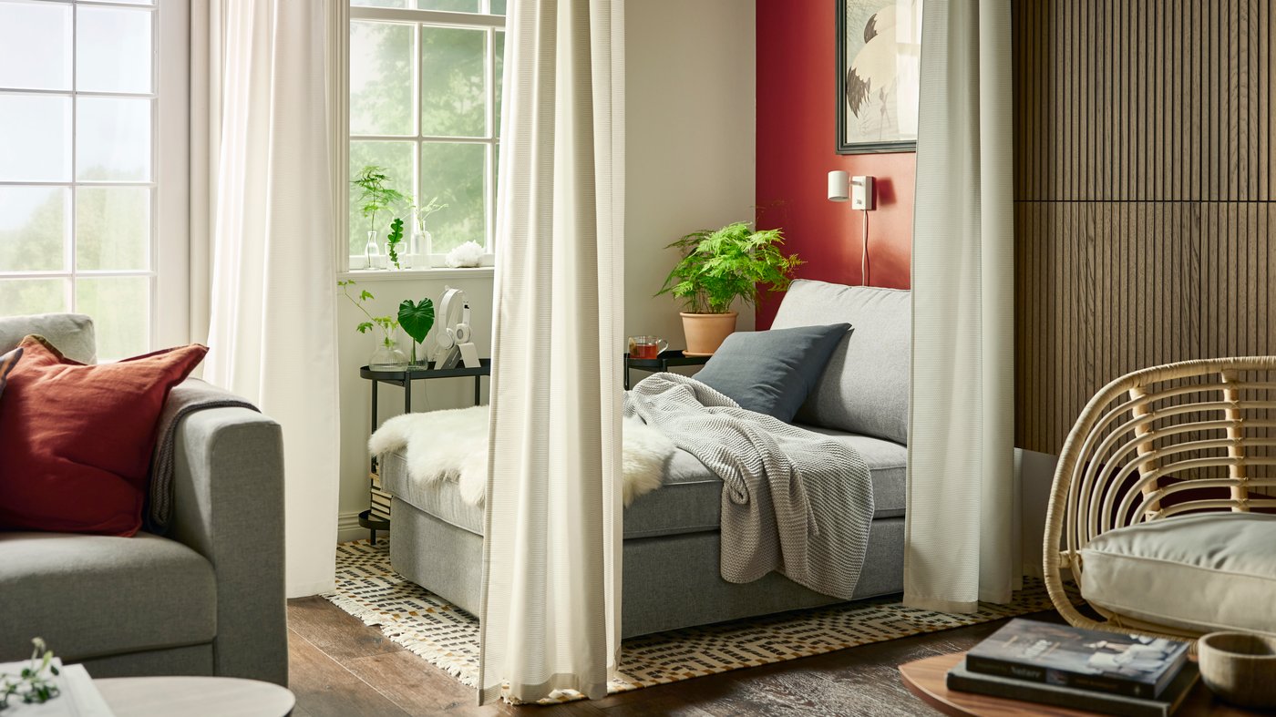 Una chaise-longue KIVIK gris amb una manta INGABRITTA de color gris clar darrere d'unes cortines insonoritzants GUNNLAUG blanques que serveixen per dividir l'espai.