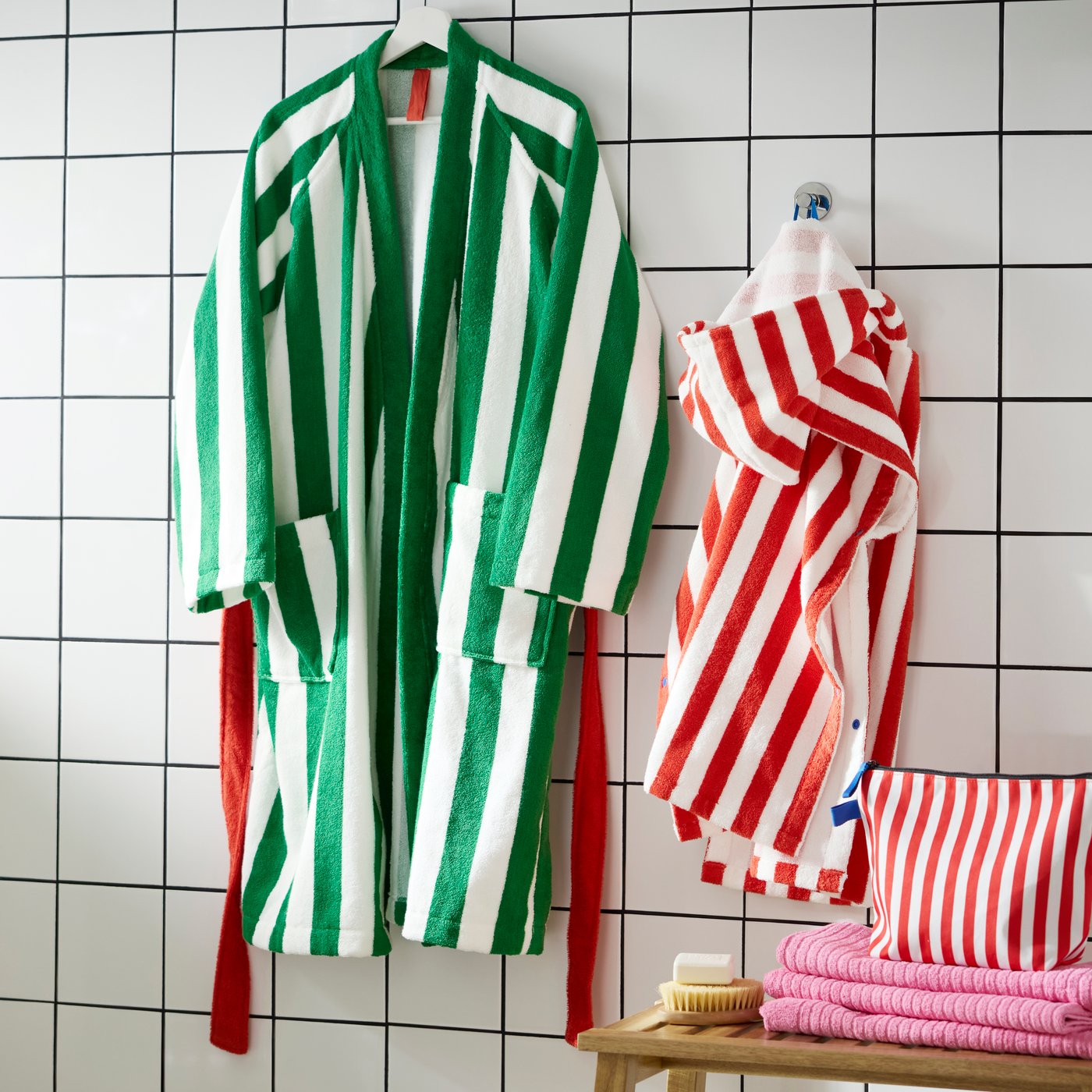 Ein großer SKOGSSALLAT-Bademantel mit grünen und weißen Streifen hängt an einer weißen Wand neben einem kleinen Poncho mit roten Streifen.