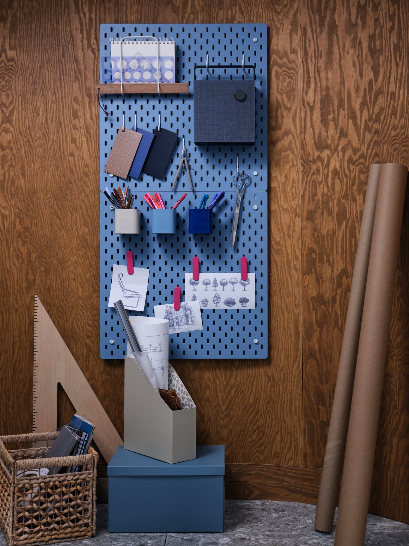 A wall with two blue SKÅDIS pegboards, SKÅDIS accessories filled with office supplies, a Bluetooth speaker and small storage.
