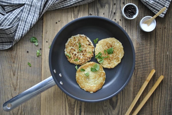 Trois galettes dorées et croustillantes, garnies d'herbes aromatiques, sont disposées dans une poêle noire sur une table en bois, à côté de laquelle se trouvent des baguettes et une nappe à carreaux.