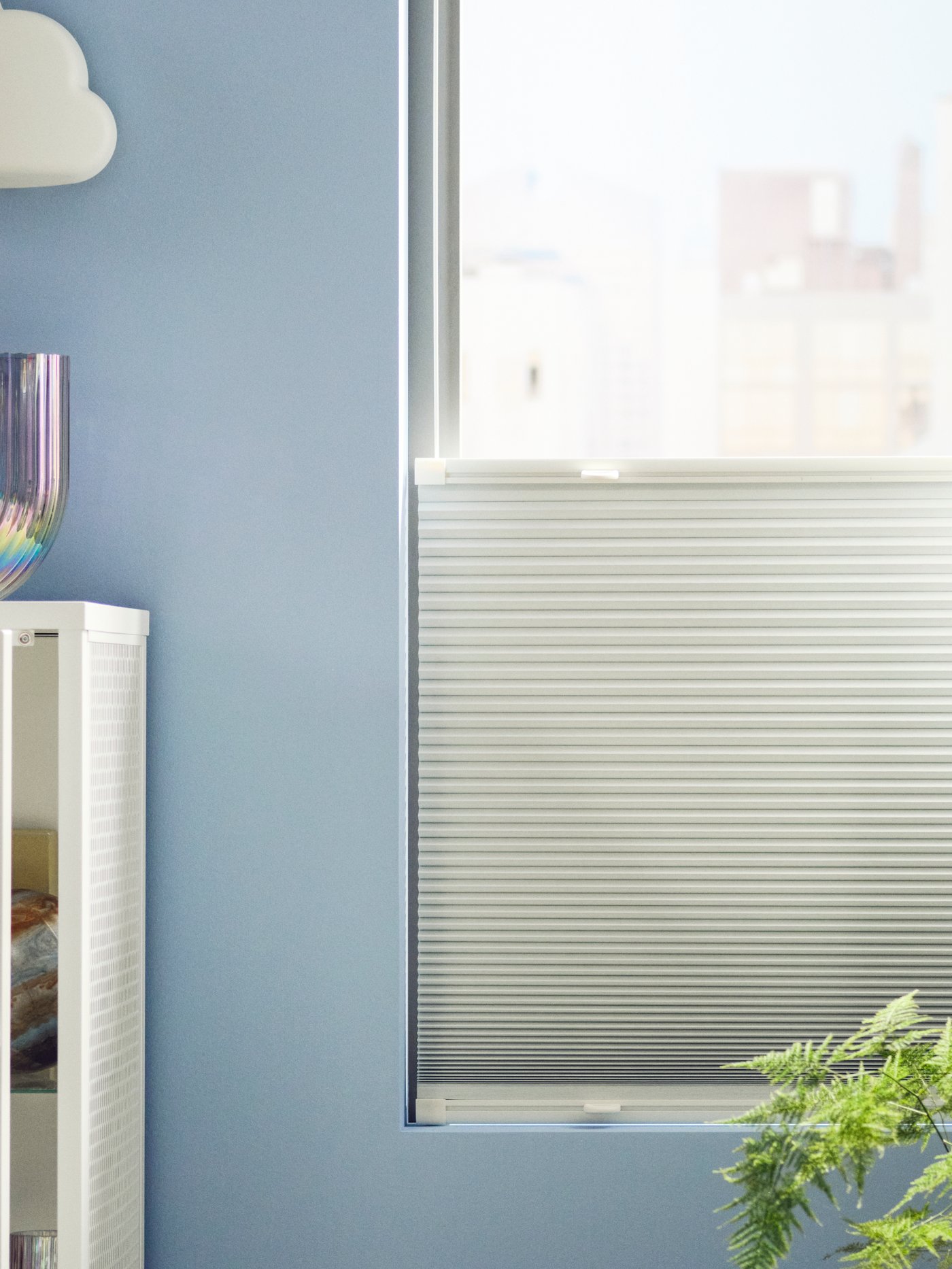 HORNVALLMO plissegardin med mørklægningseffekt er trukket halvt ned i et vindue i et rum med lyseblå vægge.