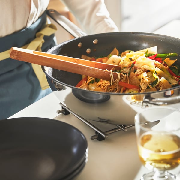 Ein IKEA 365+ Wok aus Edelstahl mit Antihaftbeschichtung wird mit Essen auf einem Tisch gebracht.