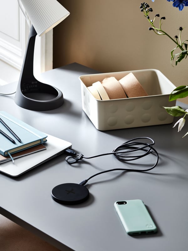 A desk with a black LIVBOJ wireless charger and some other objects on it: a smartphone, a laptop, notebooks and a desk lamp.
