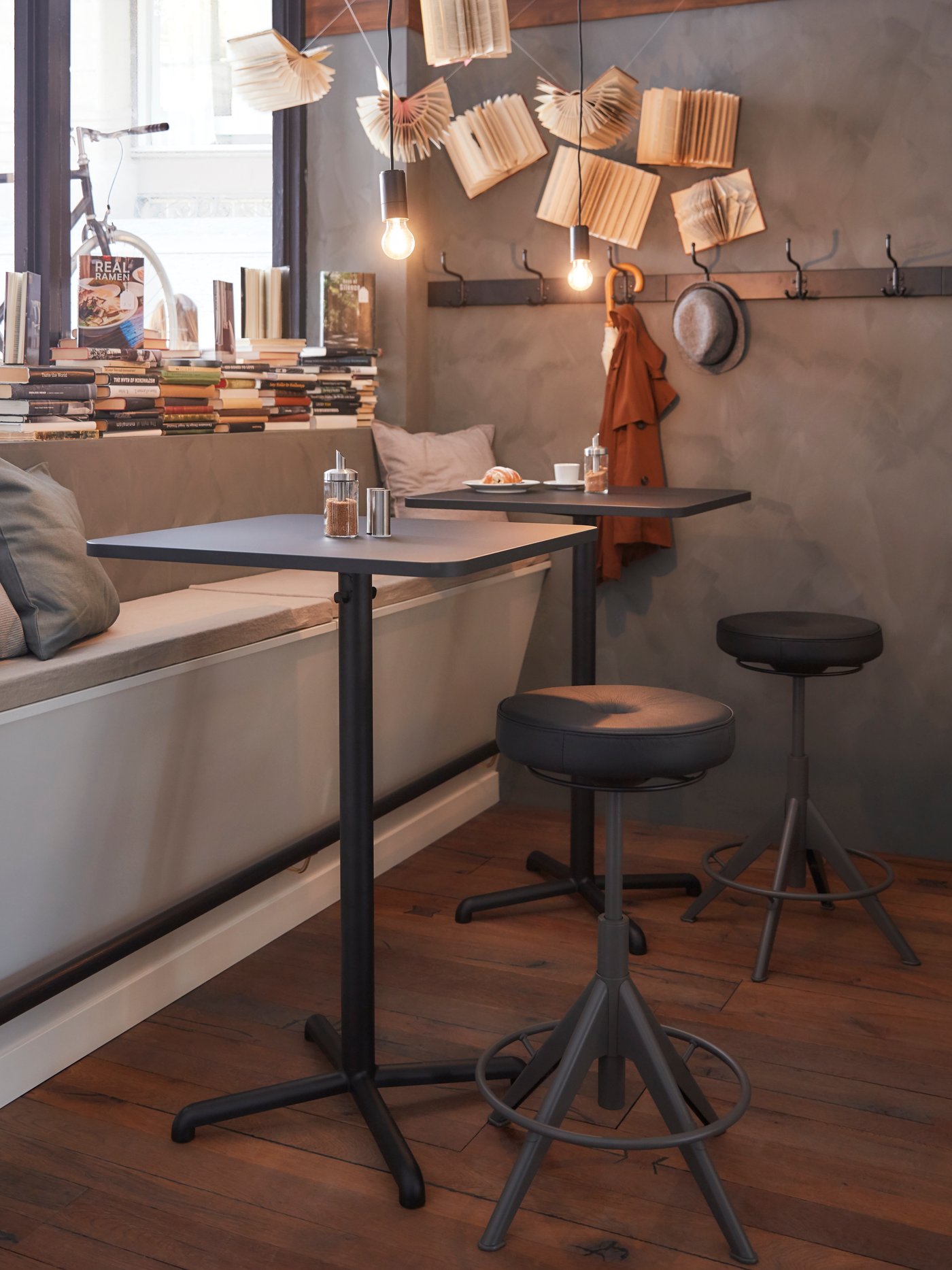 שני שולחנות מרובעים בצבע שחור לצד אדן חלון שעליו ספרים, ספסל עם כריות אפורות ושני שרפרפים שחורים לישיבה ועמידה.